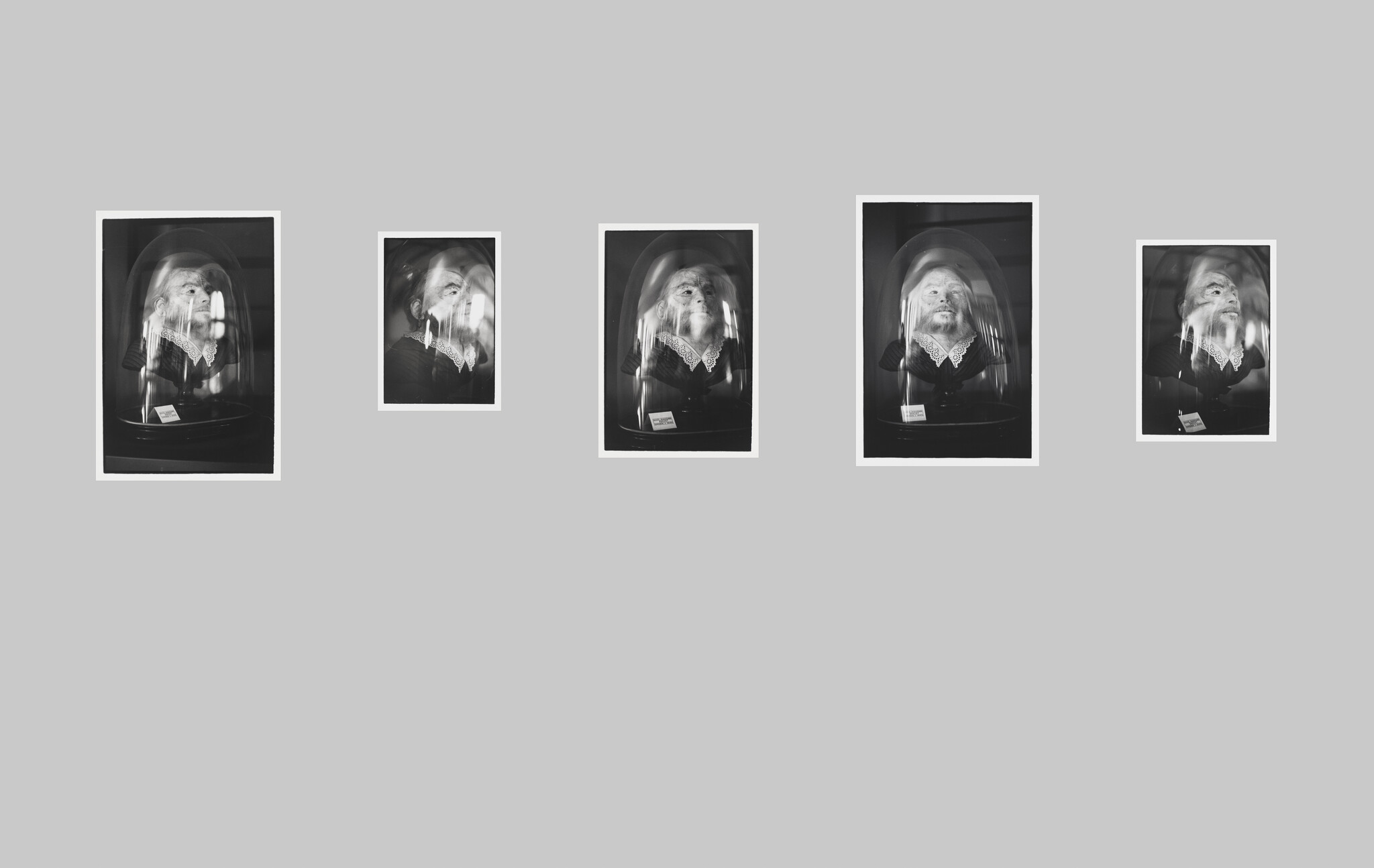 Five framed black-and-white portraits of a man with a lace collar under glass domes on a gray wall.