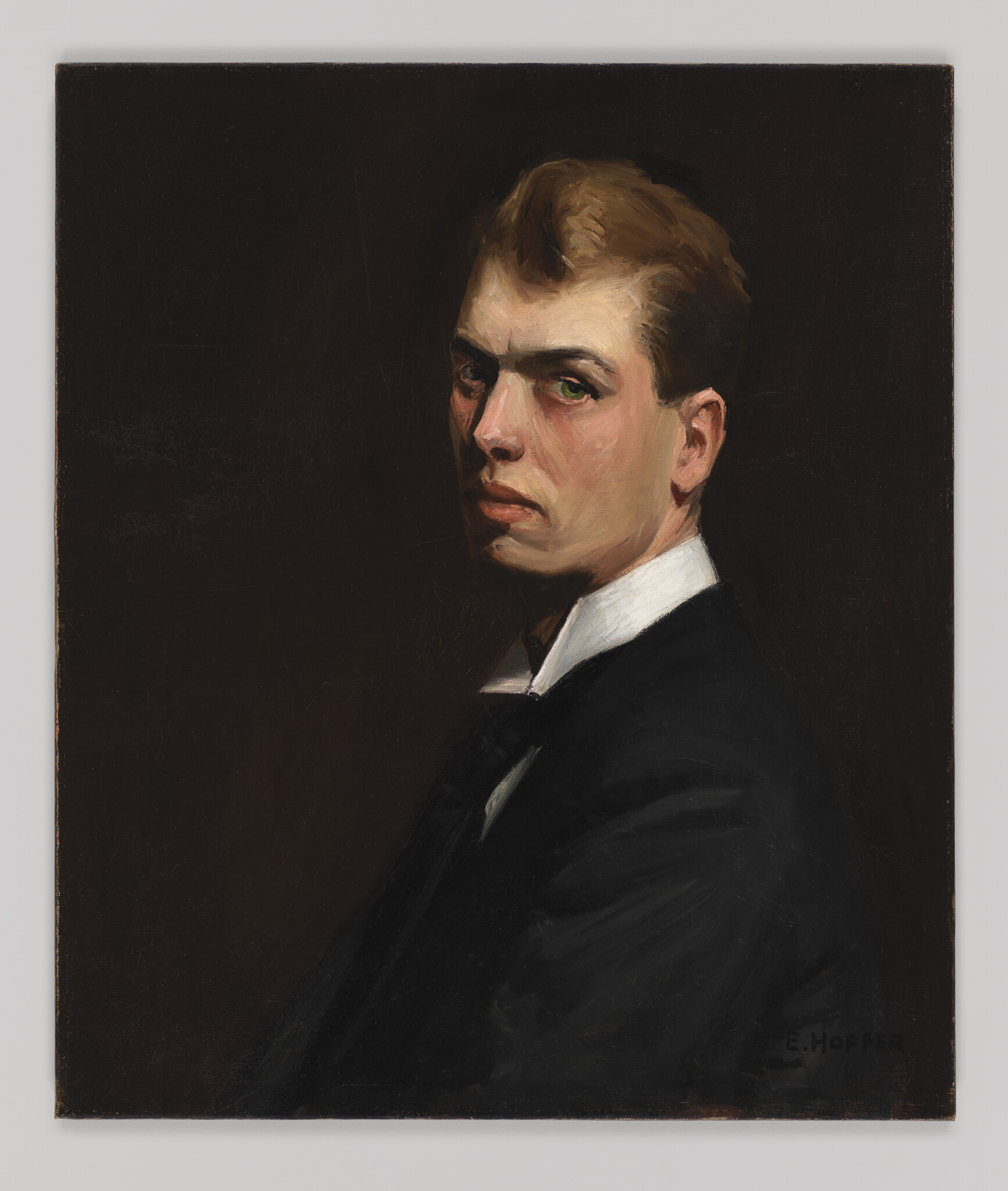 A young man in a dark suit looks over his shoulder with a serious expression.