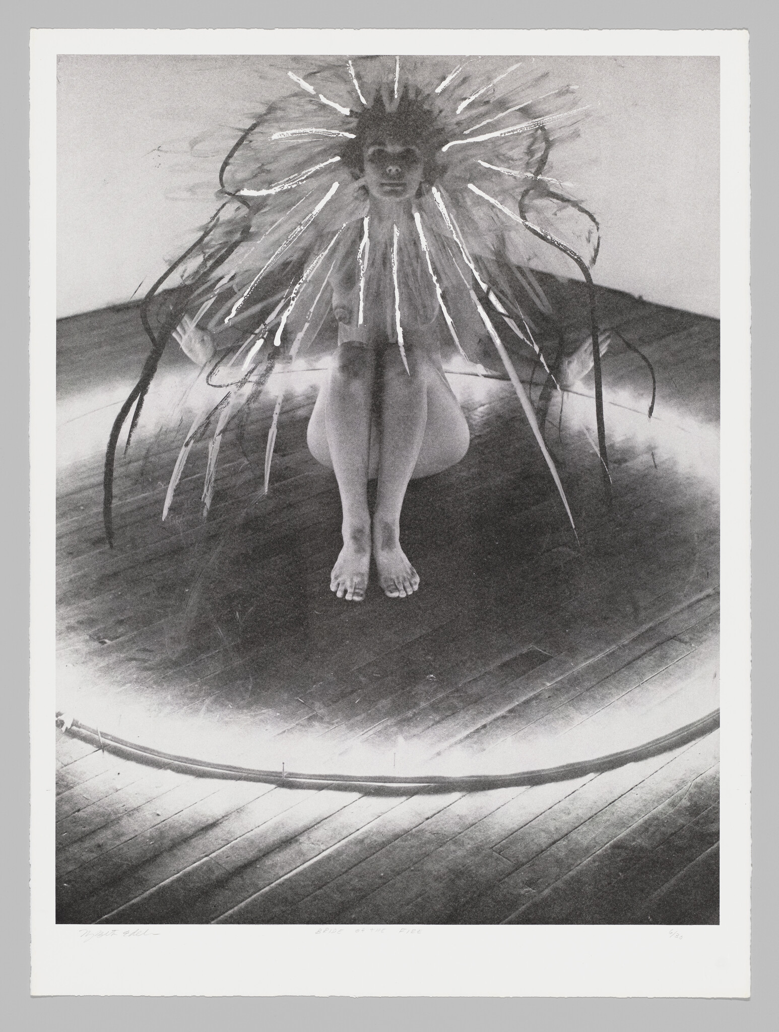 A black and white photograph depicting a person squatting on a wooden floor with a dramatic, spiky halo-like structure around their head, creating a sunburst effect. The figure appears to be part of an artistic performance or installation.