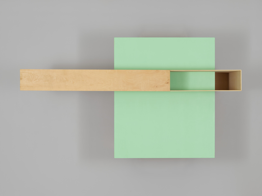 A minimalist wooden shelf with a light natural finish intersects two mint green panels mounted on a gray wall, creating a modern and simplistic aesthetic. The shelf is partially recessed into the larger panel, revealing a hollow interior.