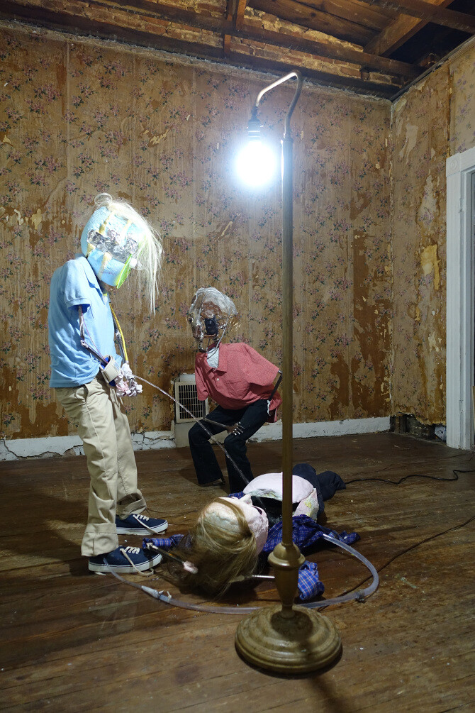 A surreal scene in a room with exposed walls and wooden floors, featuring three figures with distorted, wrapped faces resembling mummies. One figure stands upright in a blue shirt and khaki pants, another sits in a red shirt and black pants, and the third lies on the ground in a blue and white checkered shirt. A bright lamp on a stand illuminates the scene, casting shadows on the walls.