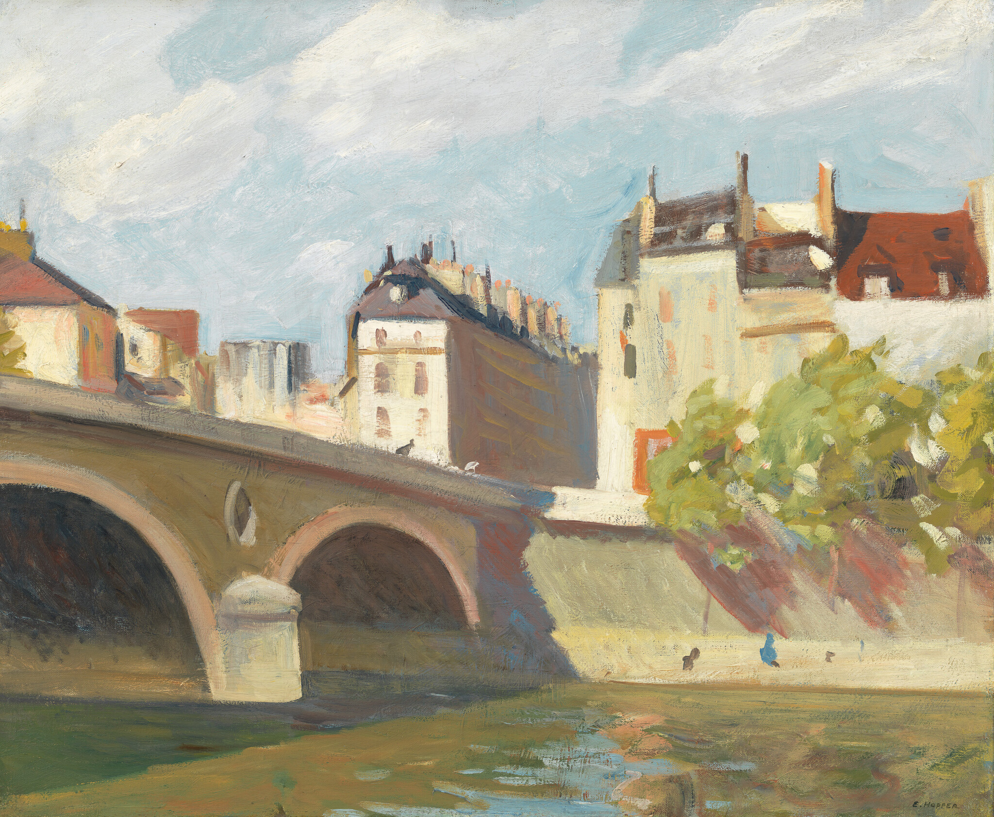 A sunlit stone bridge spans a river beside trees and buildings with people walking along the bank.