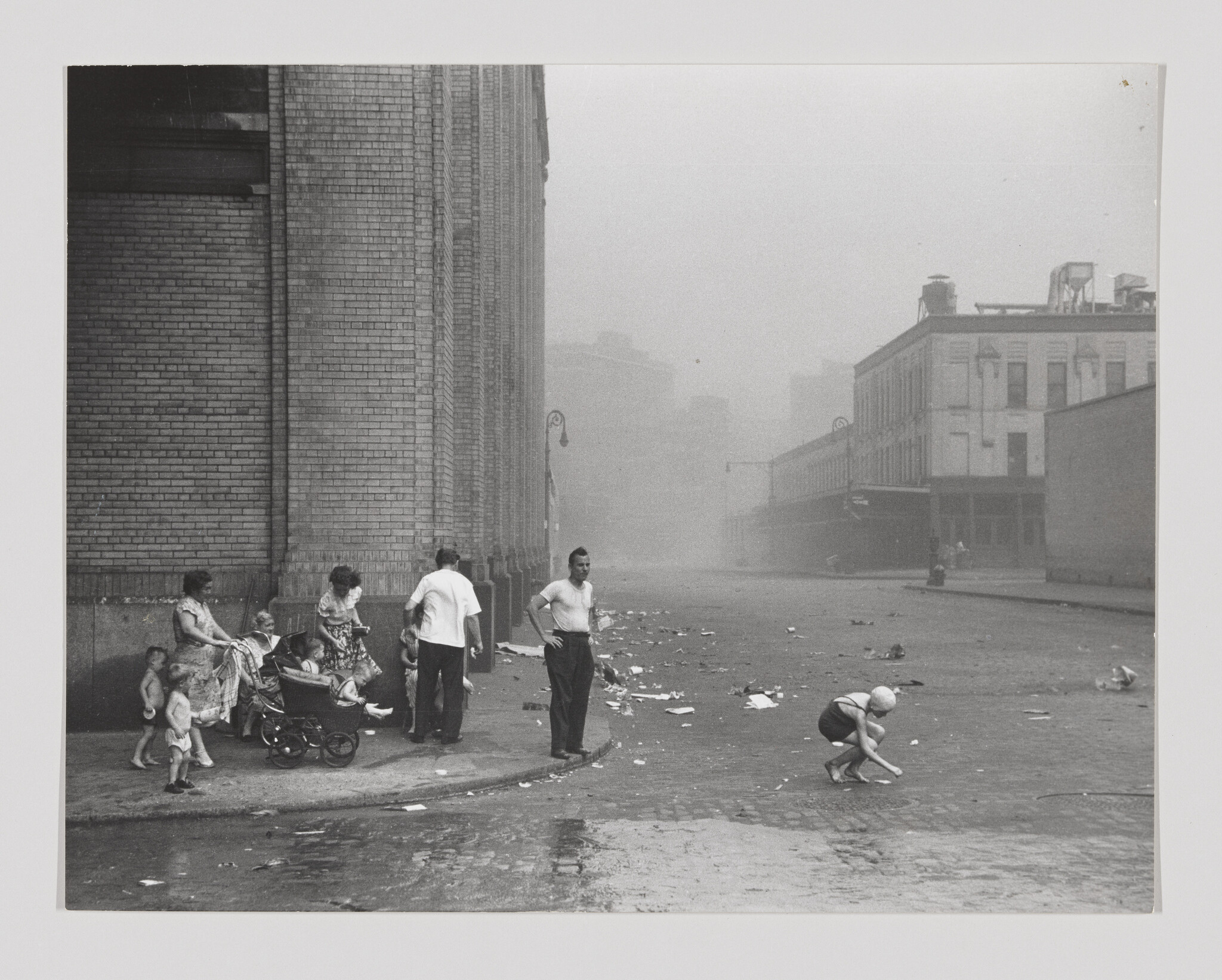 A group of city residents stand by a brick building while a child crouches picking up litter from the street.