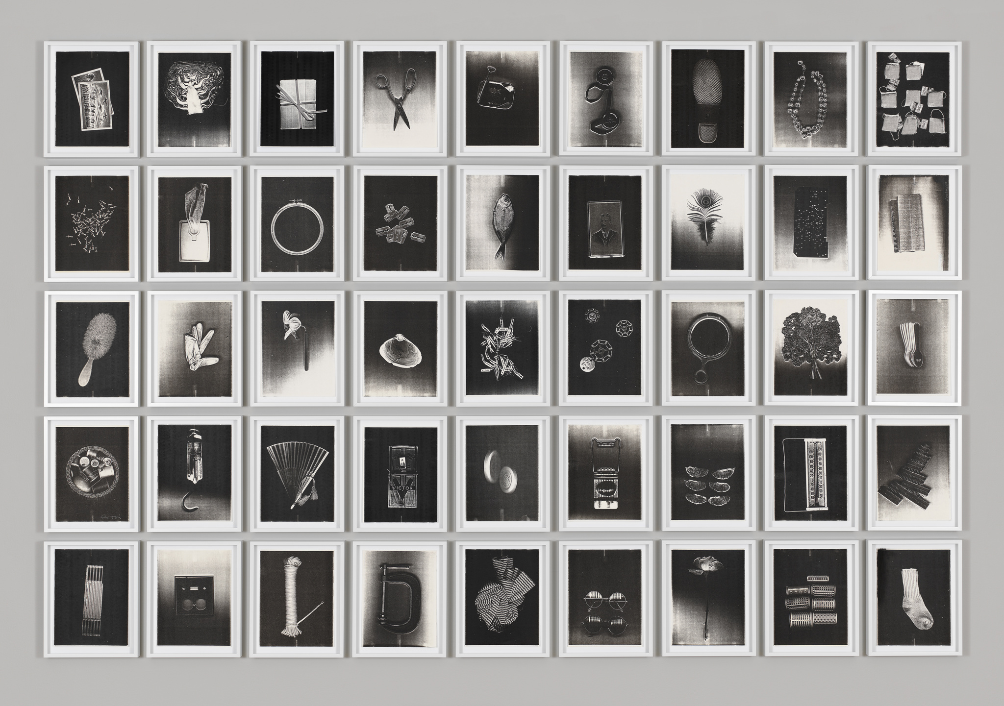 A collection of framed black and white photographs arranged in a grid on a wall, each depicting a different object or small group of objects, with a minimalist and high-contrast aesthetic.