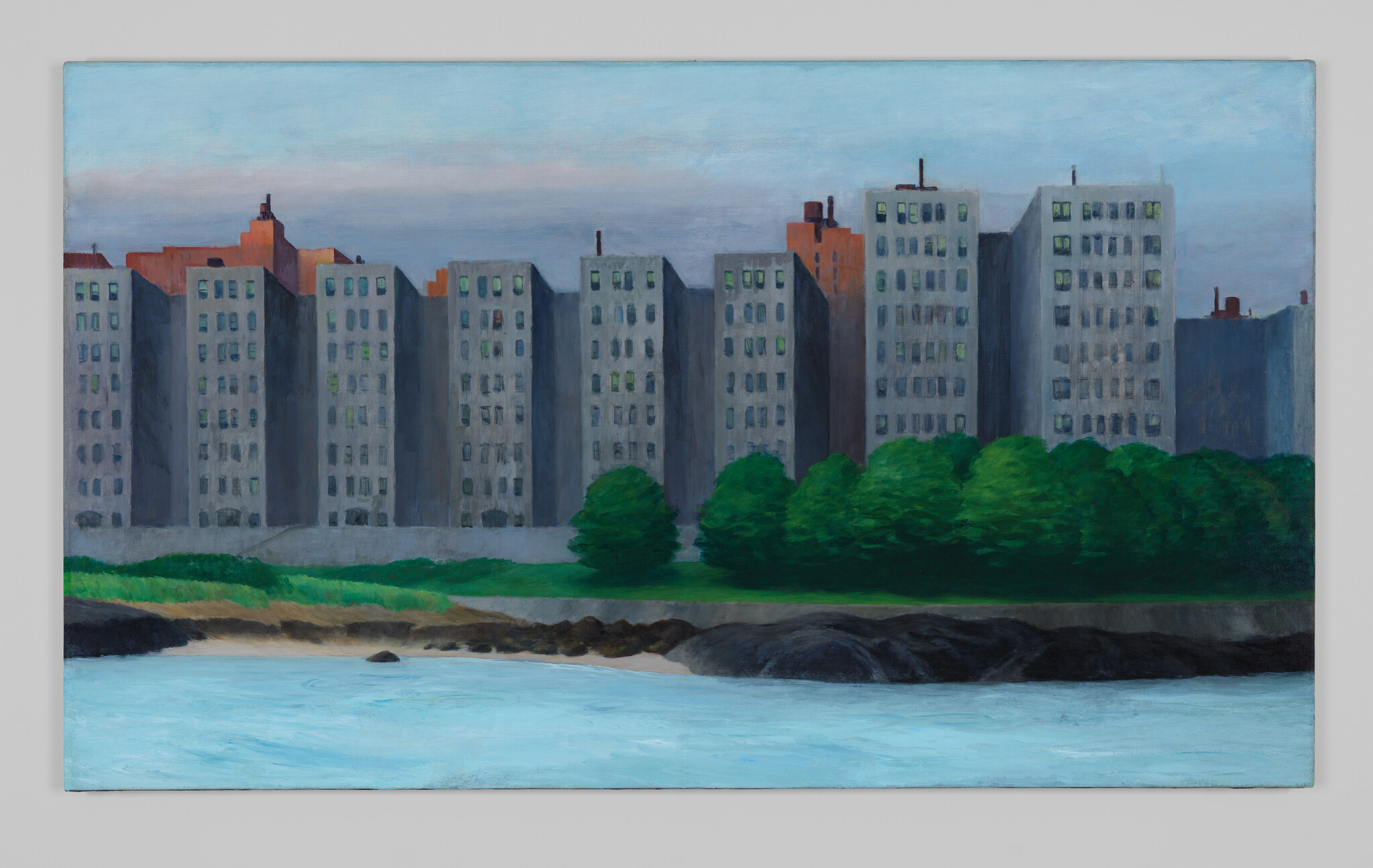 A row of gray apartment buildings stands behind green trees along a calm shoreline.