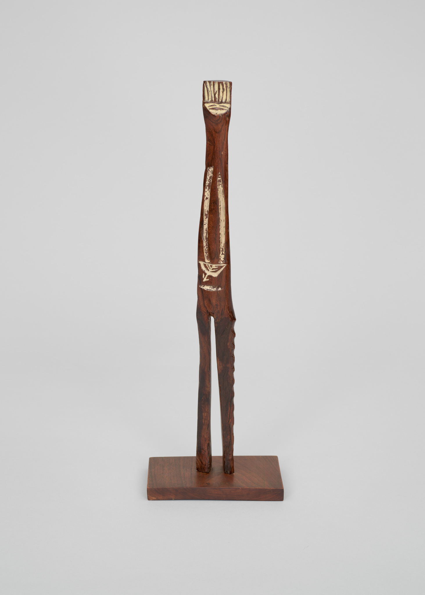 A slender, abstract wooden sculpture with a narrow opening running vertically through the center, mounted on a flat wooden base against a plain background. The top of the sculpture is carved to resemble a stylized head or crown.