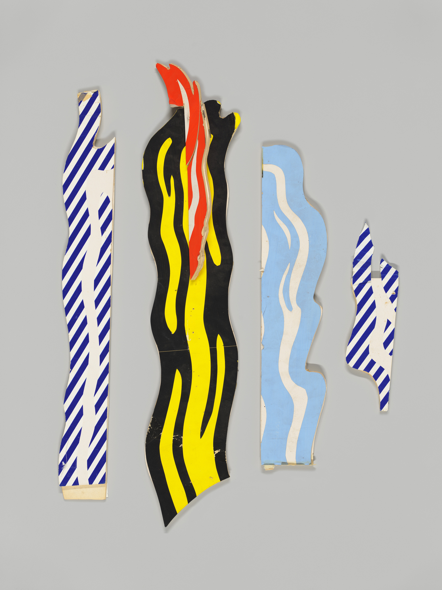 Four abstract wooden cutouts with painted designs against a gray background. From left to right: the first and fourth pieces feature blue and white diagonal stripes, the second piece has a black background with yellow and red flame-like patterns, and the third piece is painted with blue and white wavy lines.