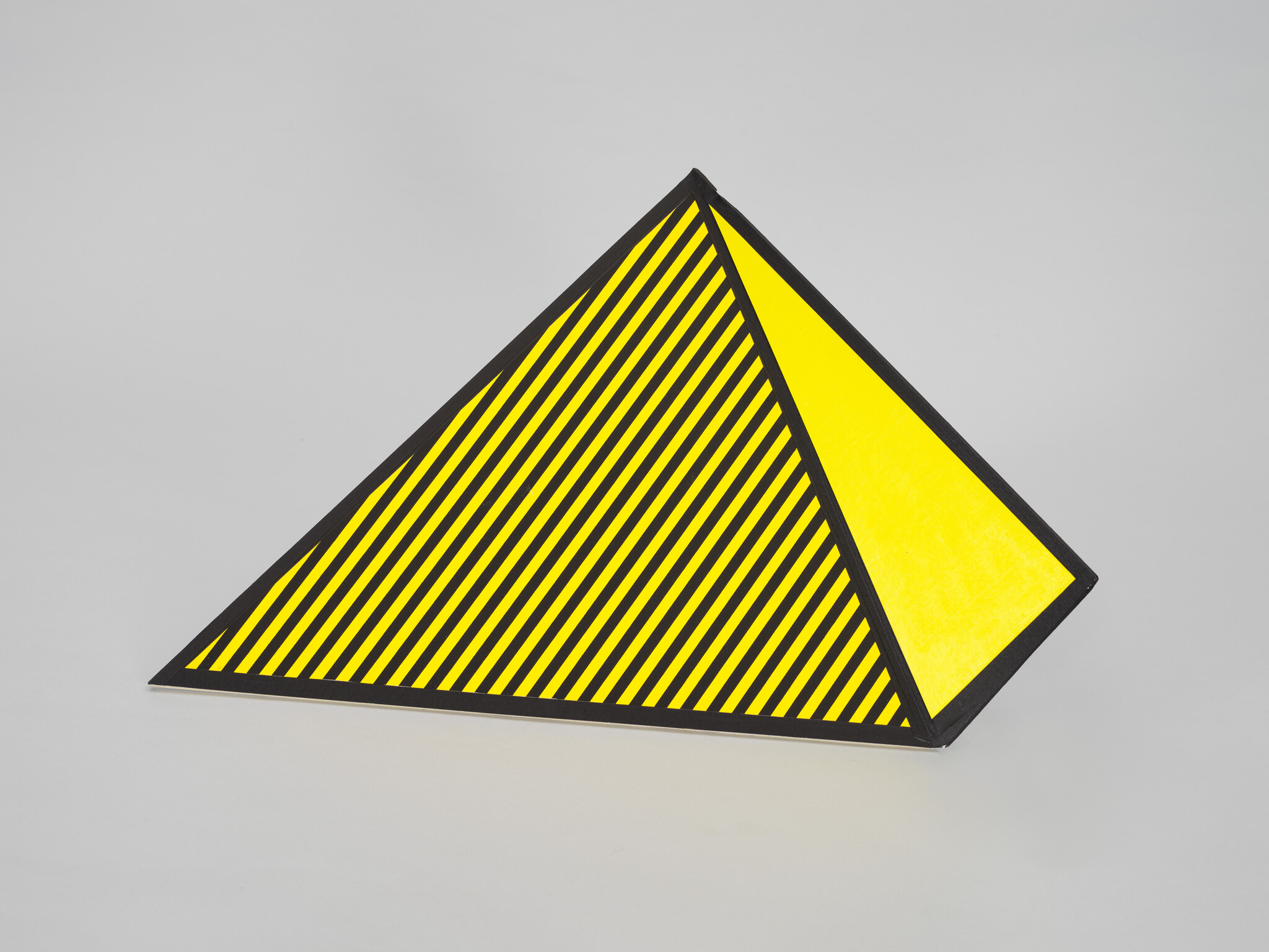A small triangular pyramid with yellow faces, one striped with diagonal black lines, on a gray surface.