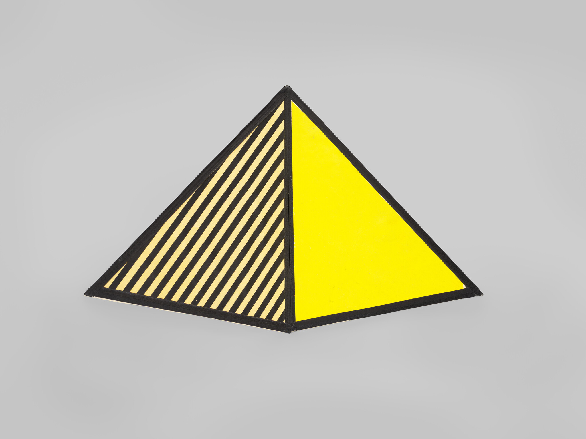 Small triangular sculpture with one solid yellow face and one yellow face with black diagonal stripes.