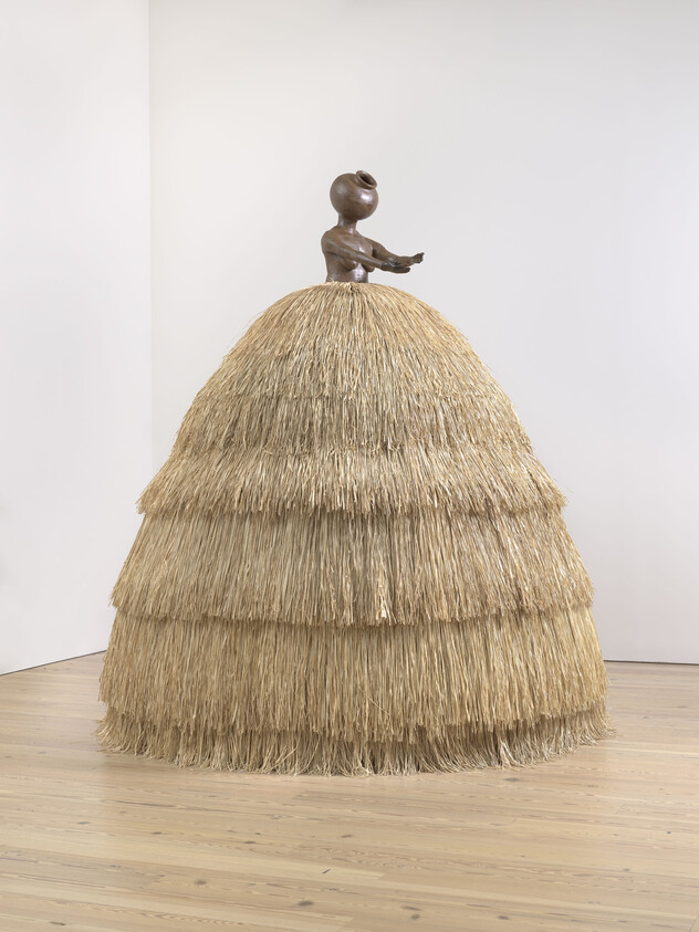 A large sculpture with a tiered straw skirt and a brown figure of a child on top, arms outstretched.