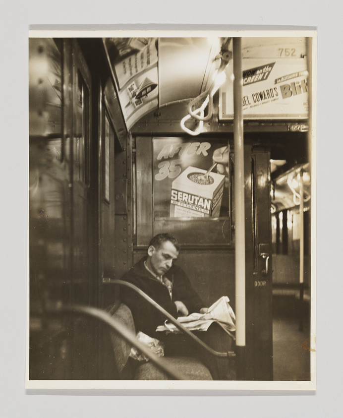 Man reading a newspaper on a vintage subway train with old advertisements overhead.