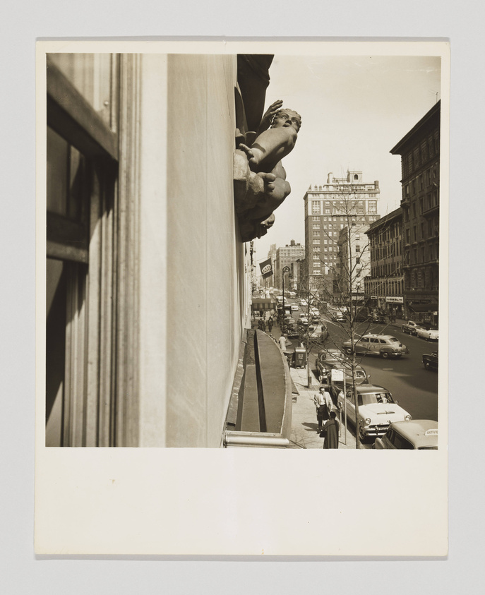 A vintage black and white photograph capturing a city street scene from an elevated angle, with the focus on a decorative stone sculpture of a human figure protruding from the corner of a building. The background shows bustling city life with cars and pedestrians on the street, indicative of mid-20th century urban life.