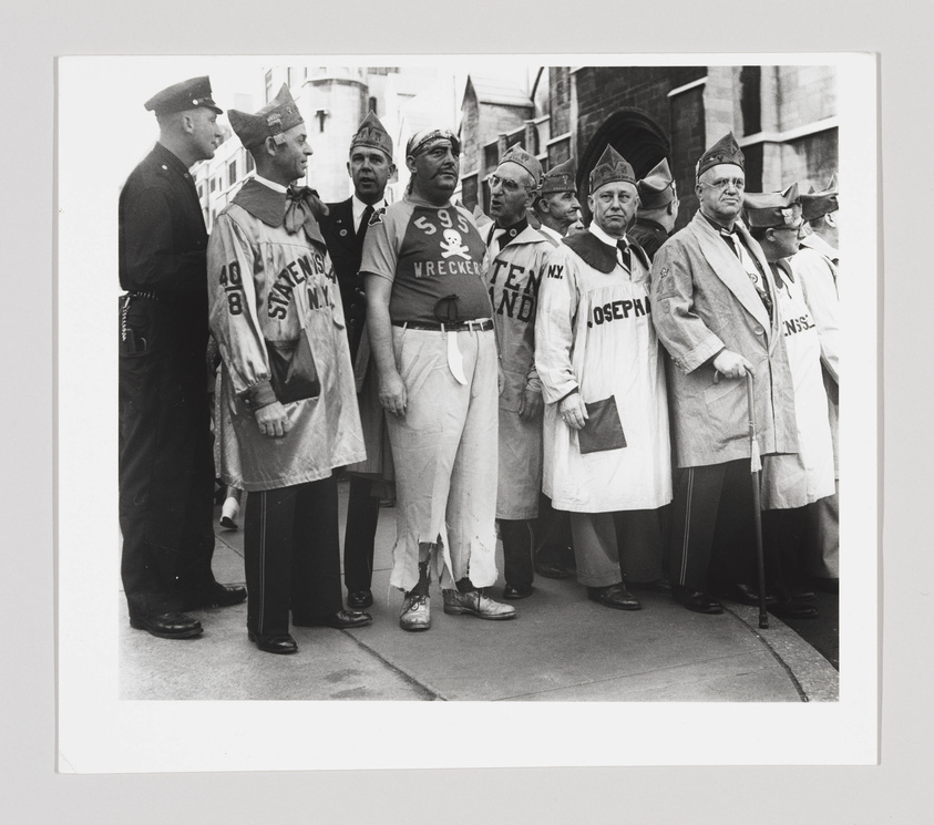 A black and white photograph showing a group of men wearing comical outfits with oversized ties and hats resembling dunce caps. They have slogans and numbers painted on their clothing, such as "59 3/4 WRECKING CREW" and "JOSEPH." Two police officers are standing to the left, conversing with one of the men in costume. The group appears to be part of a parade or public event, with a city street and buildings in the background.