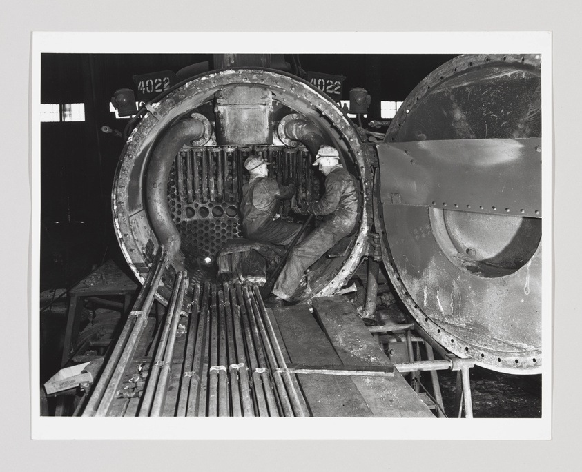 Black and white photograph of two workers repairing a large locomotive engine, with one inside the open engine compartment and the other standing outside, surrounded by tools and engine parts.