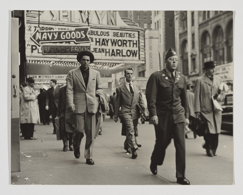 A black and white photograph capturing a bustling city street scene with pedestrians, including a prominent figure of a soldier in uniform walking towards the camera. In the background, vintage signage for a theater advertising "Navy Goods," "Fabulous Beauties," and stars like "Hayworth" and "Jean Harlow" is visible, suggesting a mid-20th-century setting.