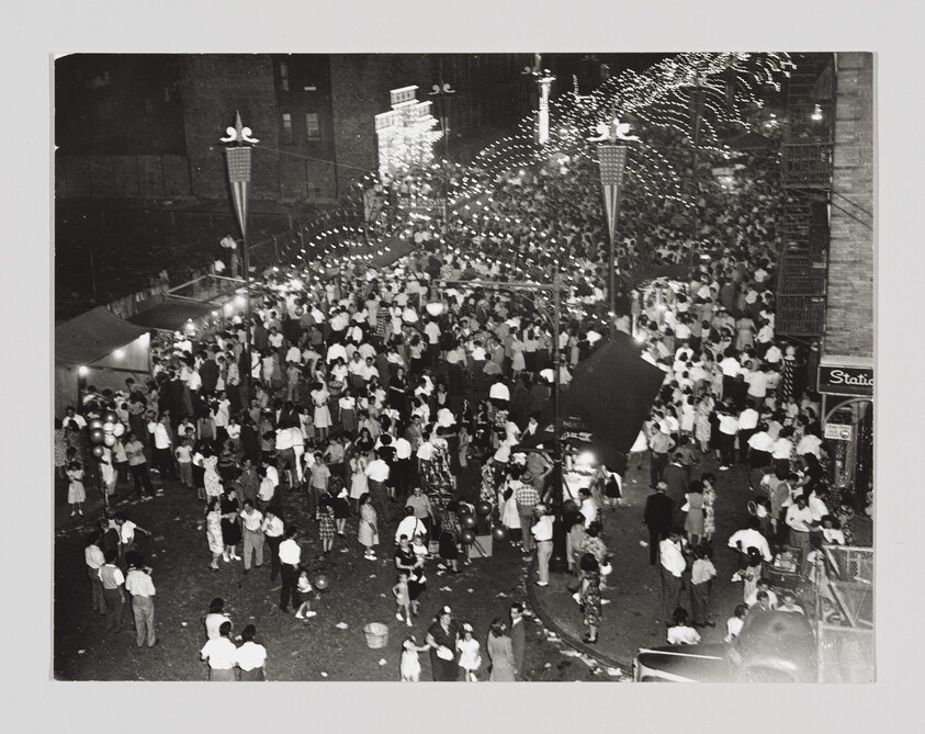 Black and white photograph capturing a bustling street festival at night. The scene is illuminated by strings of lights that create a festive atmosphere. A dense crowd of people, many dressed in light-colored clothing, fills the street, mingling, walking, and some carrying balloons. The architecture suggests an urban environment, and a sign that reads "Station" is visible in the lower right corner, indicating the proximity to a transportation hub or a themed area. The photo conveys a sense of community and celebration.