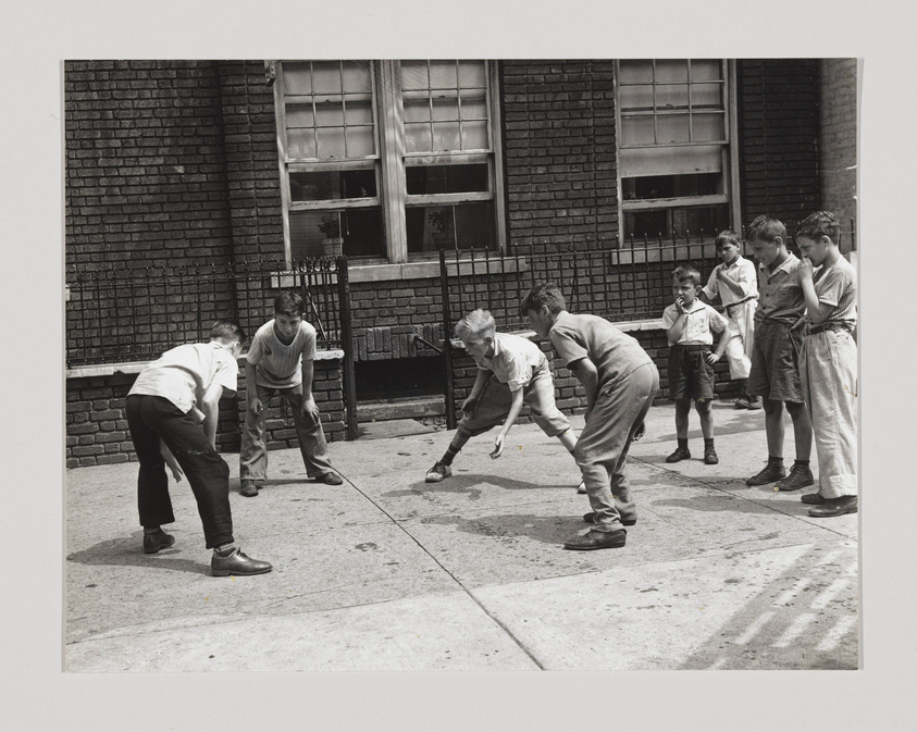 A black and white photograph of a group of boys playing a game on a city sidewalk, with two boys crouched down facing each other as if they are about to start a race, while others watch. The setting includes a brick building with windows in the background, and a metal fence to the left. The boys are dressed in casual, mid-20th-century attire.