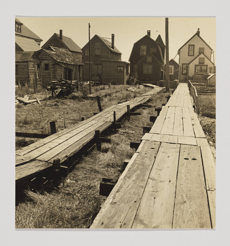 A sepia-toned photograph depicting a wooden boardwalk leading through a row of dilapidated, weathered houses. The scene is devoid of people, and the grass around the houses appears overgrown. Utility poles line the path, and the houses show signs of neglect, with peeling paint and some visible damage. The perspective is from the boardwalk itself, looking down the path between the houses.