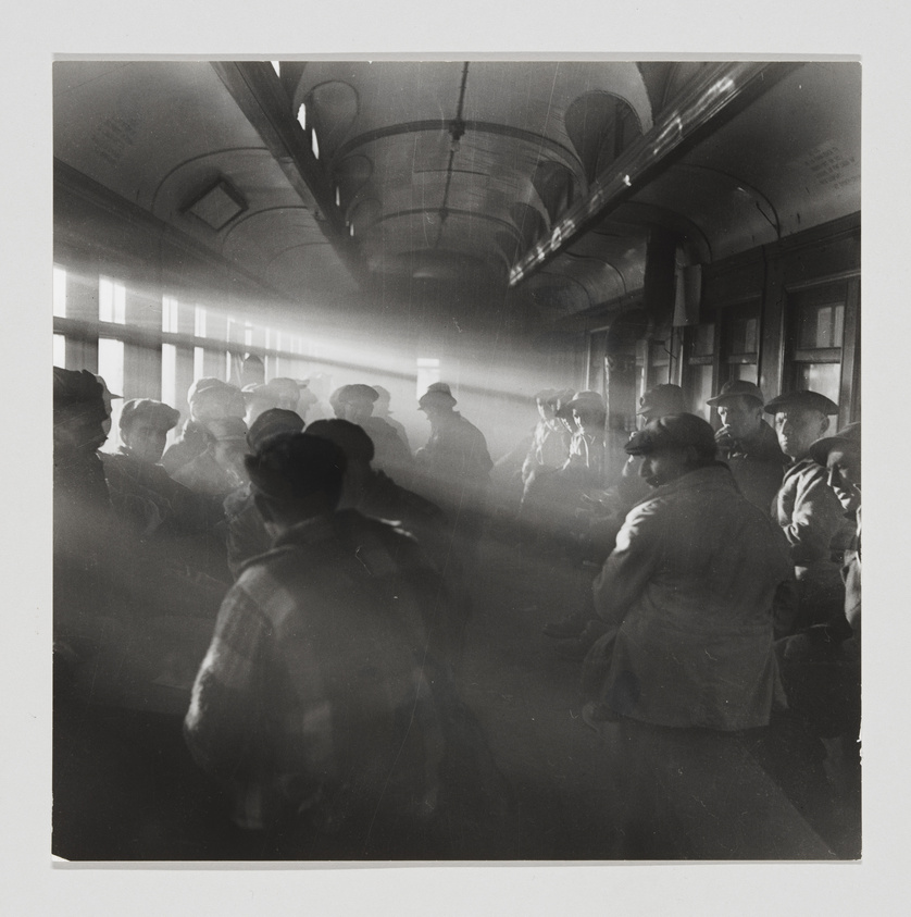 Black and white photograph capturing a group of people inside a vintage train car, with sunlight streaming through the windows creating a dramatic effect with rays of light. The passengers appear to be in casual conversation, wearing hats typical of mid-20th century fashion, and the atmosphere is filled with a hazy glow.