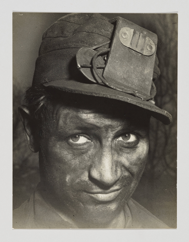 A close-up black and white photograph of a coal miner with a weathered face, his skin covered in coal dust. He is wearing a work cap with a headlamp attached, looking directly at the camera with a slight, knowing smile.