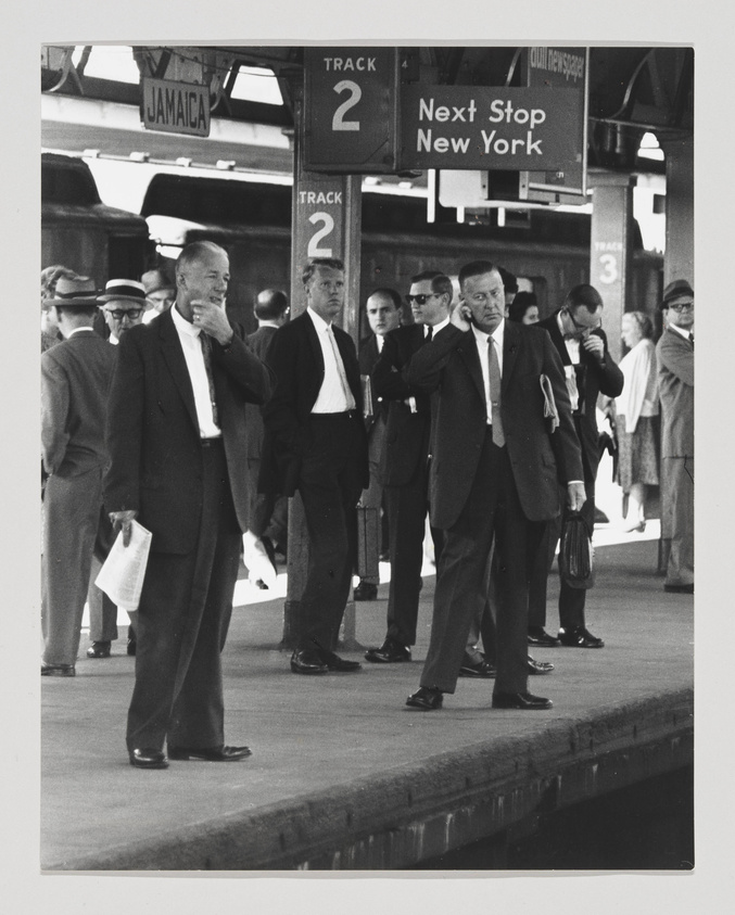 Black and white photo of commuters waiting on a train platform with signs indicating "Jamaica" and "Next Stop New York." Men in suits appear to be in mid-conversation or deep in thought, with one man prominently placing his hand on his chin.