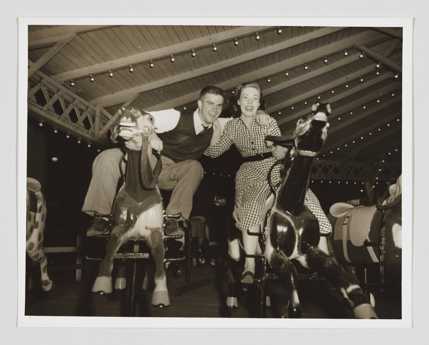 A black and white photo of a smiling man and woman riding mechanical horses on a carousel, with the man in a vest and the woman in a checkered dress, both looking joyfully at the camera. The setting appears to be an indoor amusement area with a wooden ceiling and string lights.