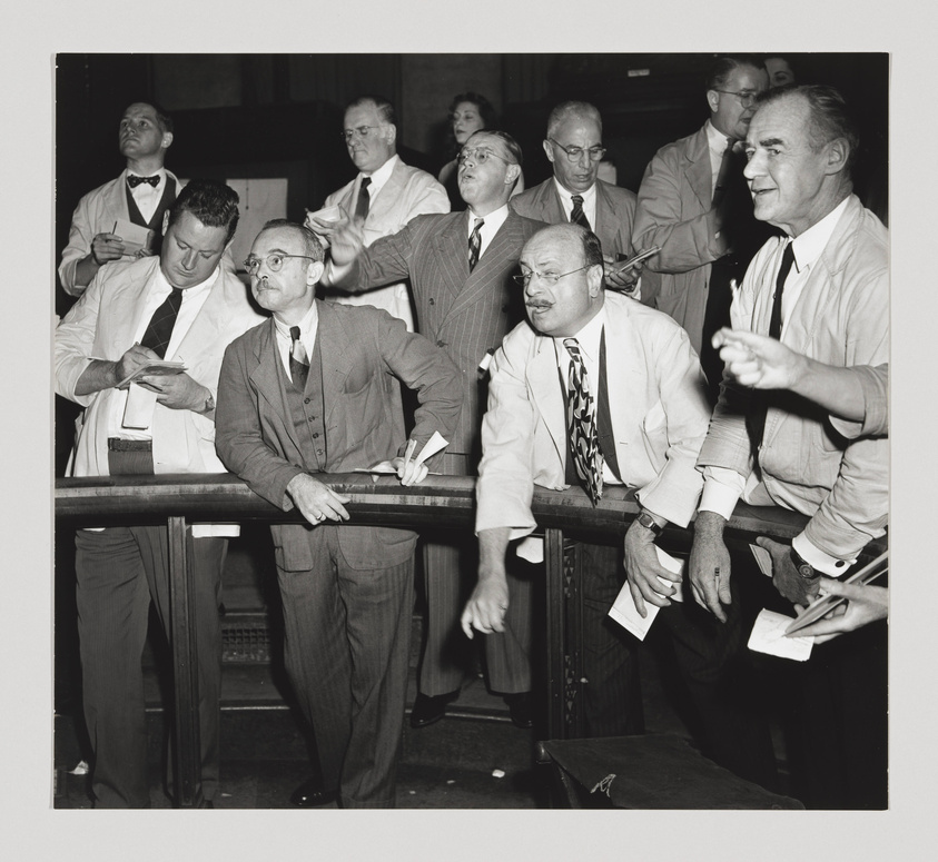 A black and white photograph of a group of men in business attire, some with pens and papers in hand, standing and sitting around a railing, seemingly focused on an event or activity not visible in the image. The setting appears to be indoors, possibly a stock exchange or betting venue, given the intense expressions and the papers that could represent tickets or financial documents.