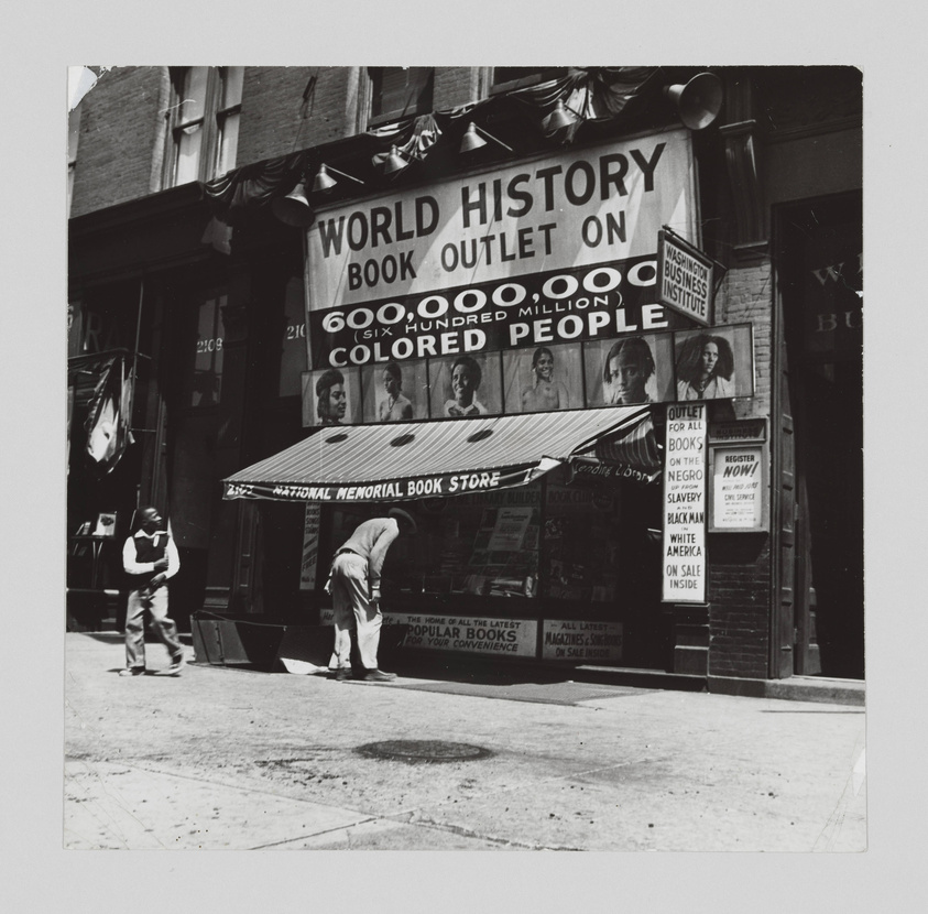 A black and white photograph of the exterior of the National Memorial Book Store. The storefront features a large sign reading "WORLD HISTORY BOOK OUTLET ON 600,000,000 (SIX HUNDRED MILLION) COLORED PEOPLE" and other signs advertising books on Negro history, slavery, and Black America. Two individuals are visible on the sidewalk, one walking by and another bending over near the entrance.