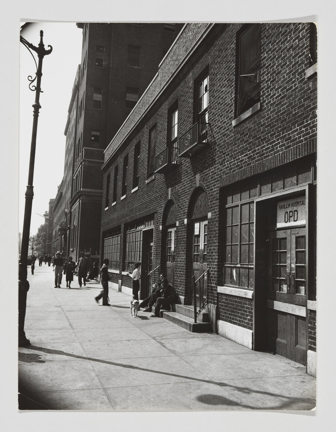 Black and white photograph of a city street scene with people walking and sitting outside a brick building labeled "Harlem Hospital OPD." A street lamp is visible on the left, and the shadows suggest it's a sunny day.