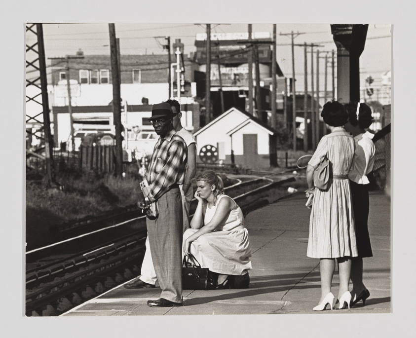 Black and white photograph of people waiting at a train station. A man in a hat and sunglasses stands with a camera, a woman sits on her suitcase looking pensive, and two other women stand in conversation, one holding a purse. Industrial buildings and power lines are visible in the background.