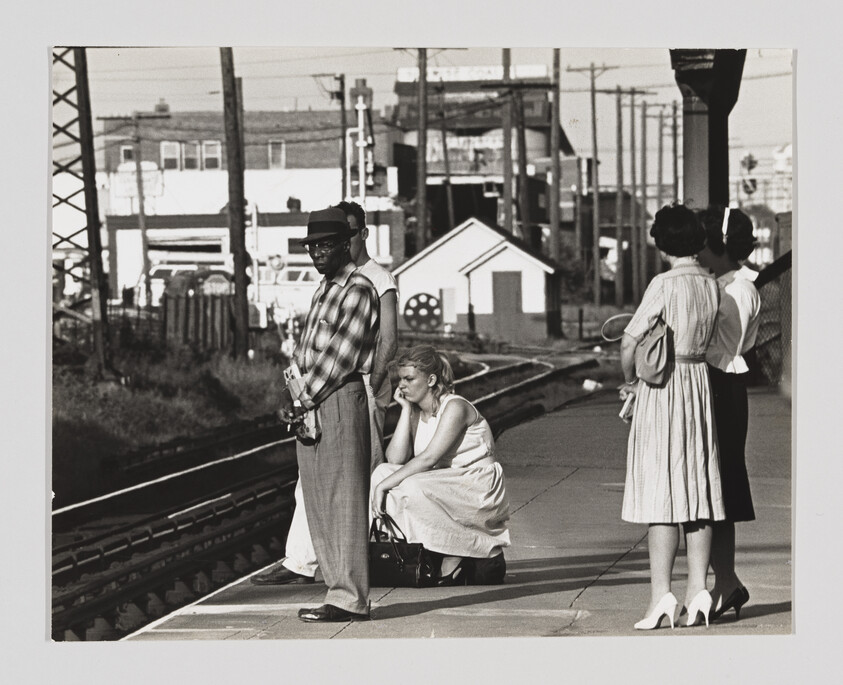 Black and white photograph of people waiting at a train station. A man in a hat and sunglasses stands with a camera, a woman sits on her suitcase looking pensive, and two other women stand in conversation, one holding a purse. Industrial buildings and power lines are visible in the background.