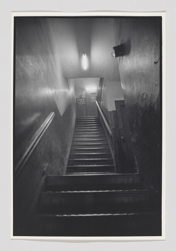 A black and white photograph depicting a dimly lit staircase leading upwards, with handrails on either side and a sign at the top that appears to read "ROSS." The walls show signs of wear and there's a light fixture on the right wall casting a glow on the steps.