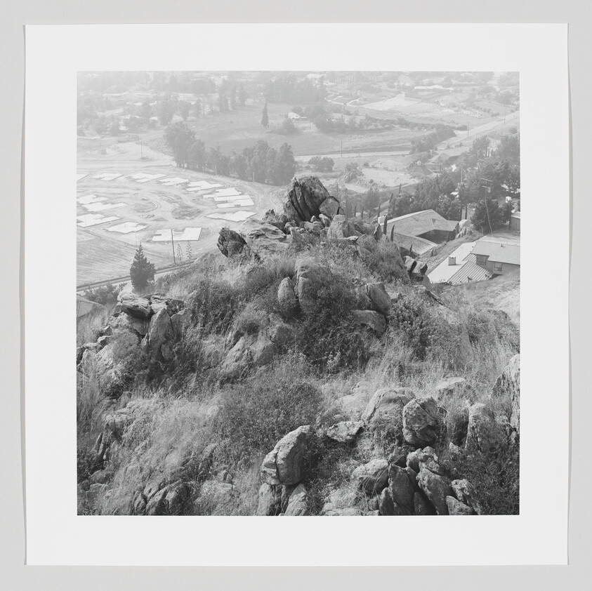 Rocky hill with grasses and boulders overlooking a suburban neighborhood and roads below.