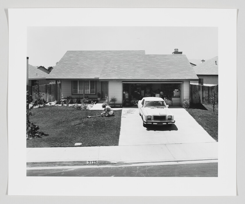 A person kneels on a front lawn tending the grass while a car sits in the driveway.