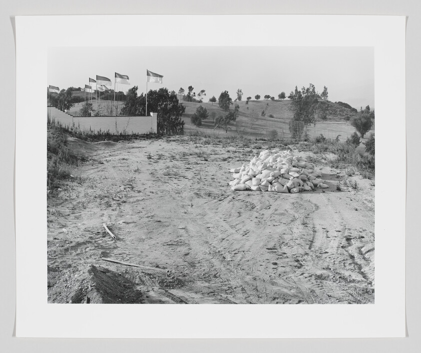 A dirt clearing with a large pile of sandbags and flagpoles near a low wall on a hill.
