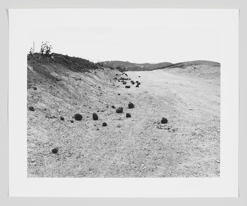 A dry dirt road strewn with scattered round rocks leading toward low, rolling hills.