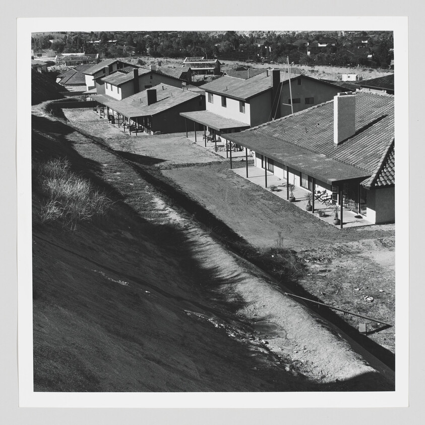 Row of single-story houses with covered porches lining a dirt path next to a steep slope.