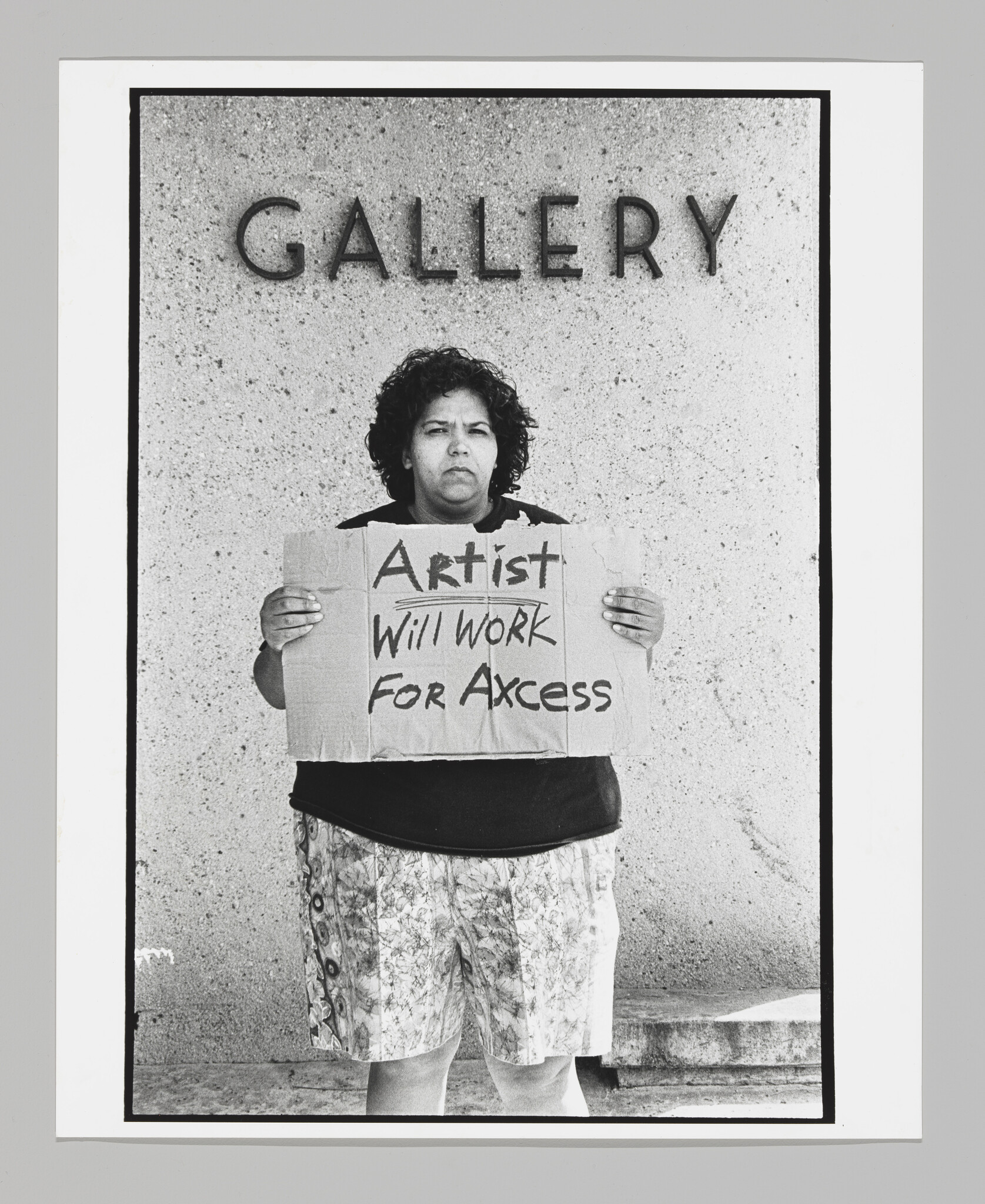 Standing in front of sign that says "Gallery," a large Latinx woman holds a cardboard sign that reads "Artist Will Work for Acess." The last word is mispelled.