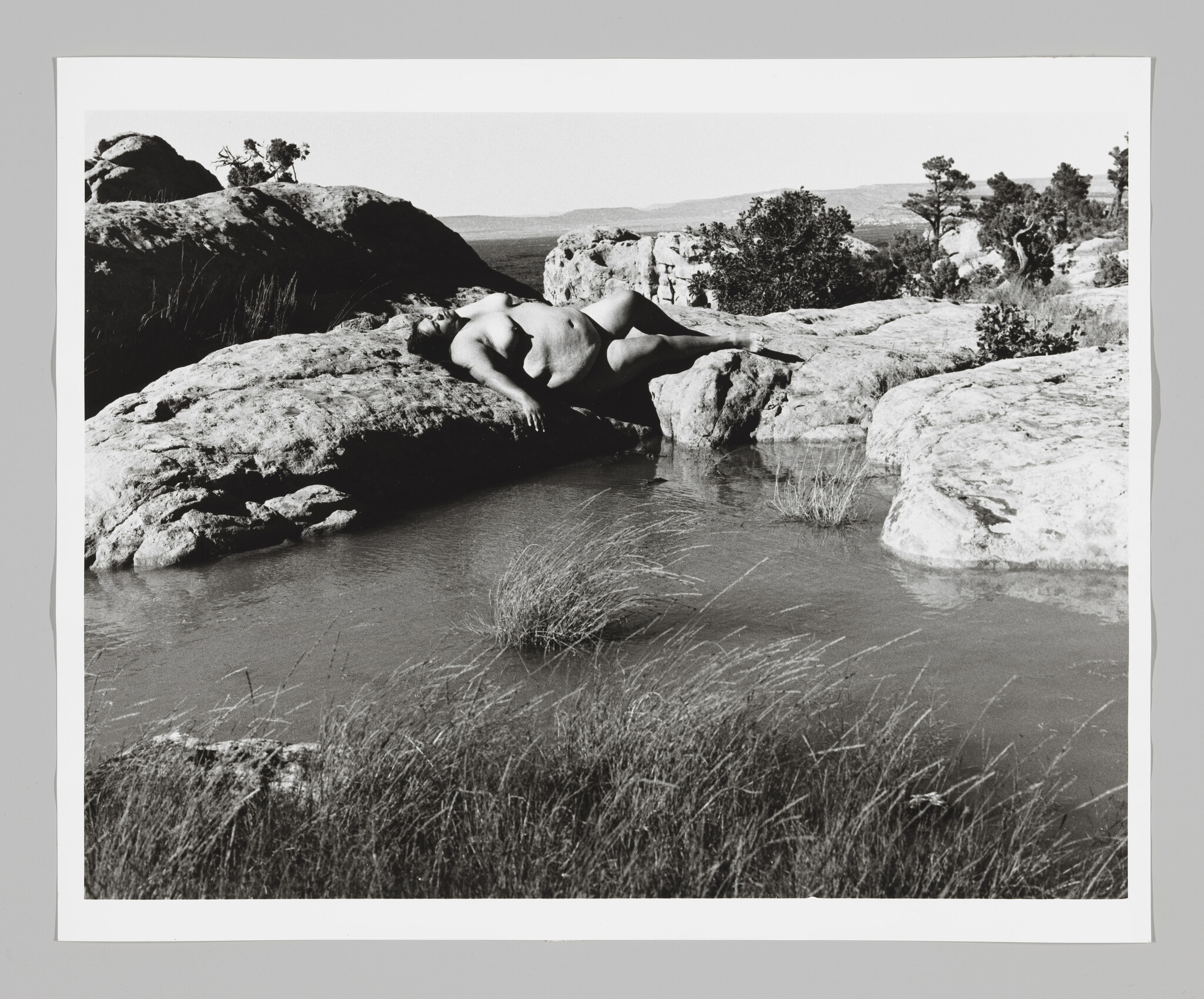 A large, naked woman reclines serenely at water's edge in a rocky landscape