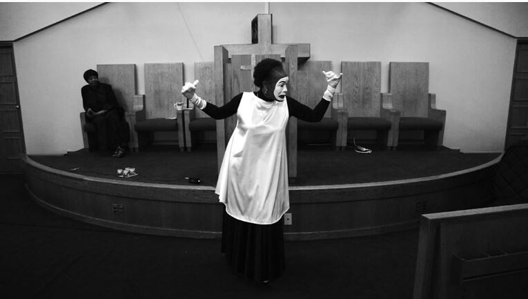 A mime-like performer in church gestures dramatically in front of a wooden cross and empty chairs.