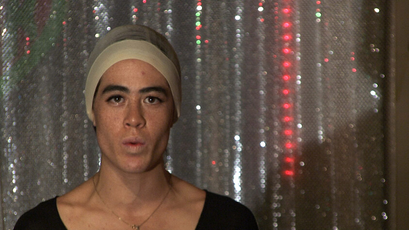 Person wearing a beige cap and black shirt stands in front of a sparkly silver curtain, looking forward.