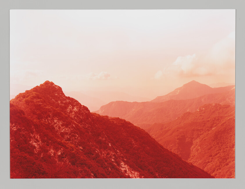 A reddish tinted landscape showing a series of mountain ridges receding into the distance under a hazy sky.