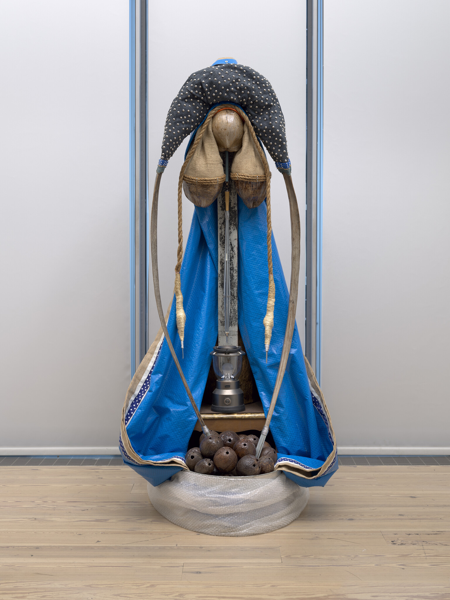 A standing mixed-media sculpture with a blue draped cloth, wooden headpiece, hanging ropes, and coconuts.