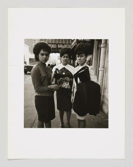 A group of three Puerto Rican women with volumnouis hair stand on a street and frown