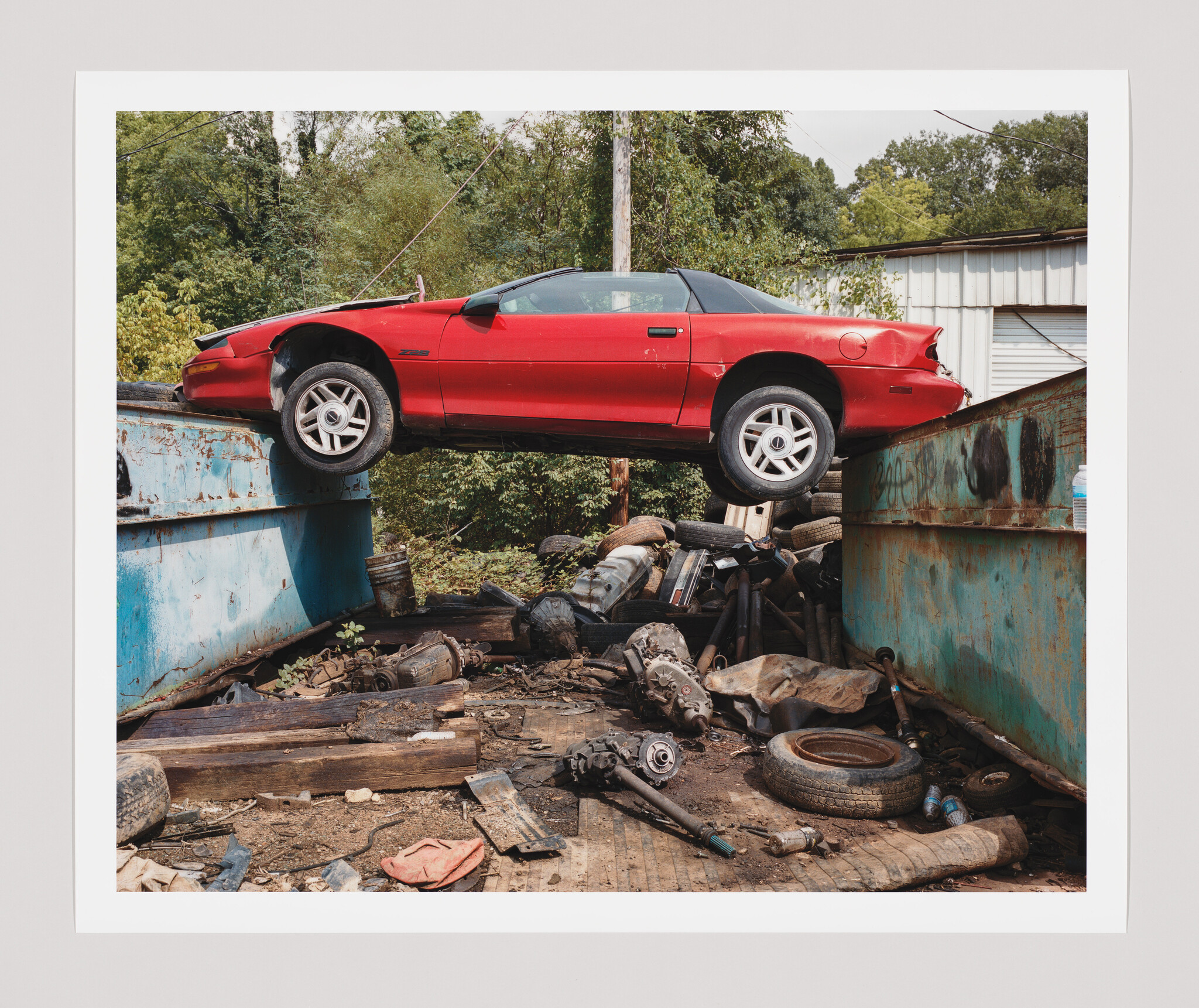 A red sports car is suspended across two rusty dumpsters above a pile of scrap parts.