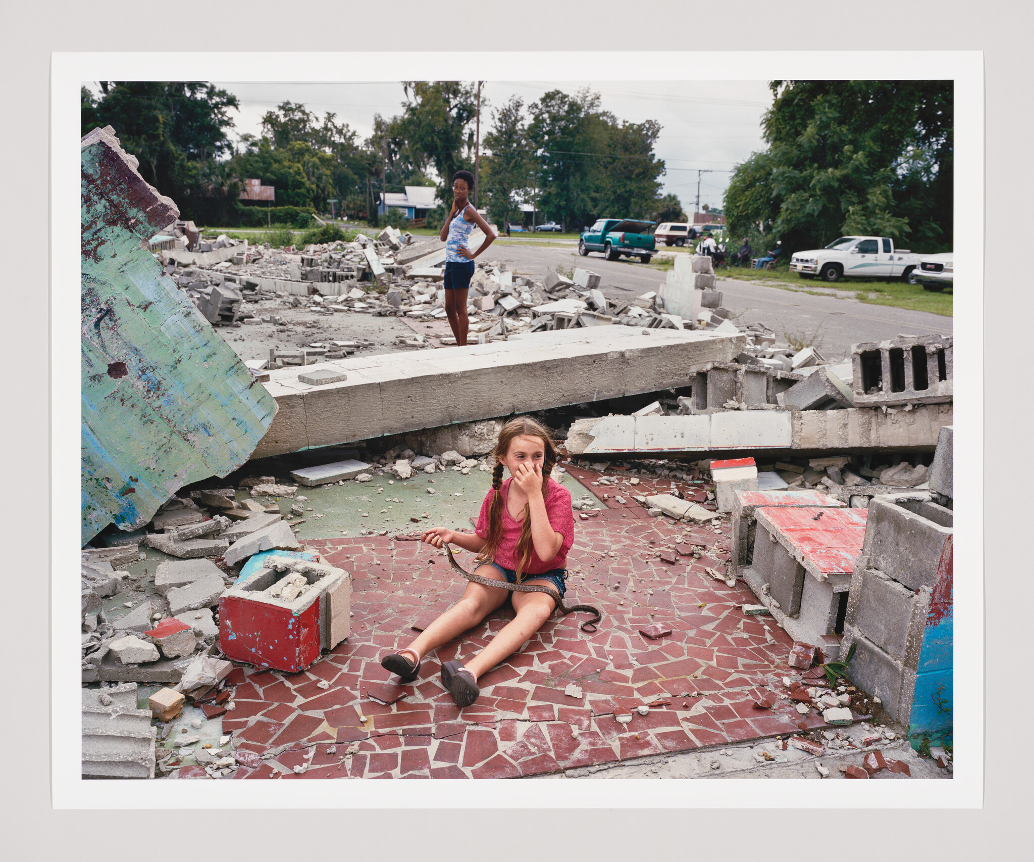 A young girl sits among rubble holding a small snake while covering her nose.