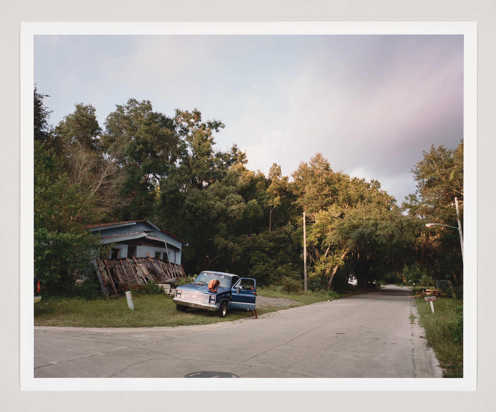 A blue pickup truck is parked by a rundown house while a person leans into its open hood.