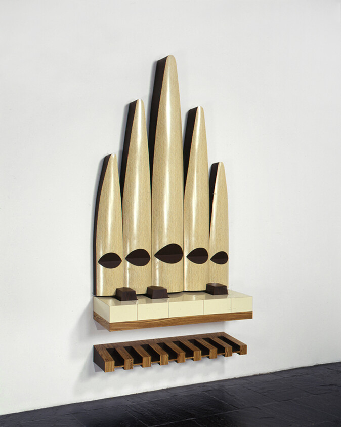 A wooden wall-mounted sculpture resembling five tall tapered organ pipes with a slatted bench shelf below.