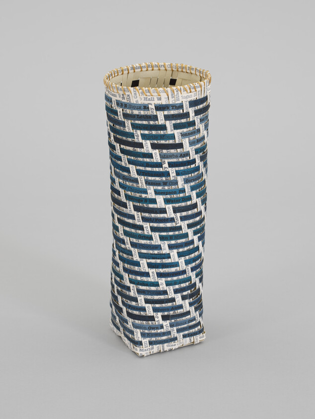 Tall woven cylindrical basket made from blue and white newspaper strips standing upright.