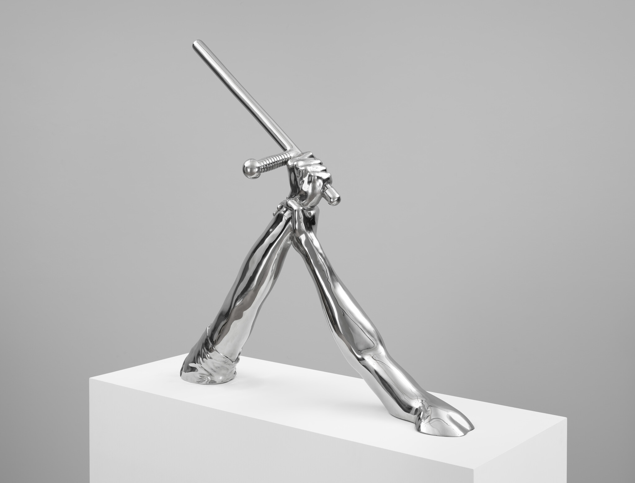 Shiny silver sculpture of two legs and hands holding a sword, standing on a white pedestal.